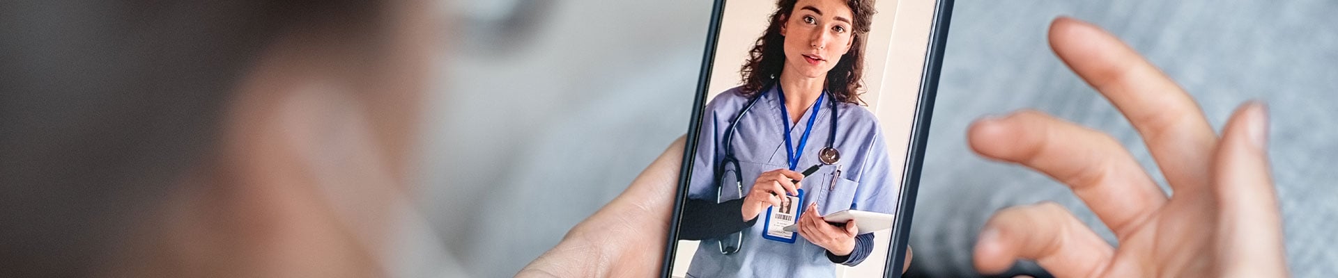 Mulher sentada e desfocada em vídeo chamada num smartphone. O smartphone está focado, onde se vê uma médica no ecrã