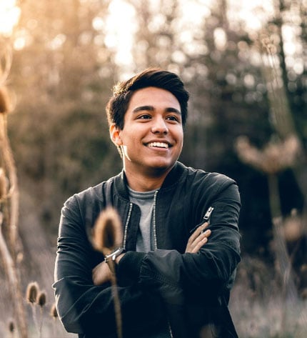 Jovem sorridente de braços cruzados numa mata ao pôr do sol