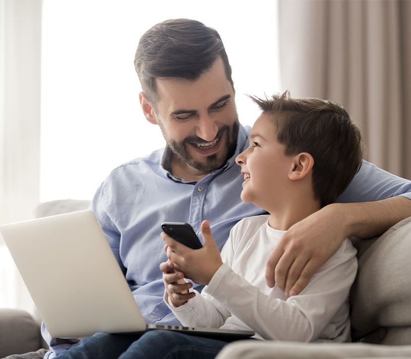 Pai sentado no sofá com portátil no colo e com o filho ao lado sorridente a brincar no telemóvel