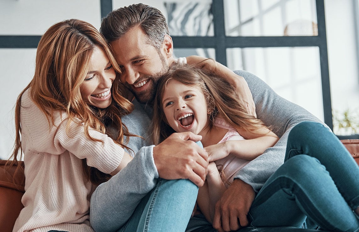Homem, mulher e filha sorridentes abraçados no sofá