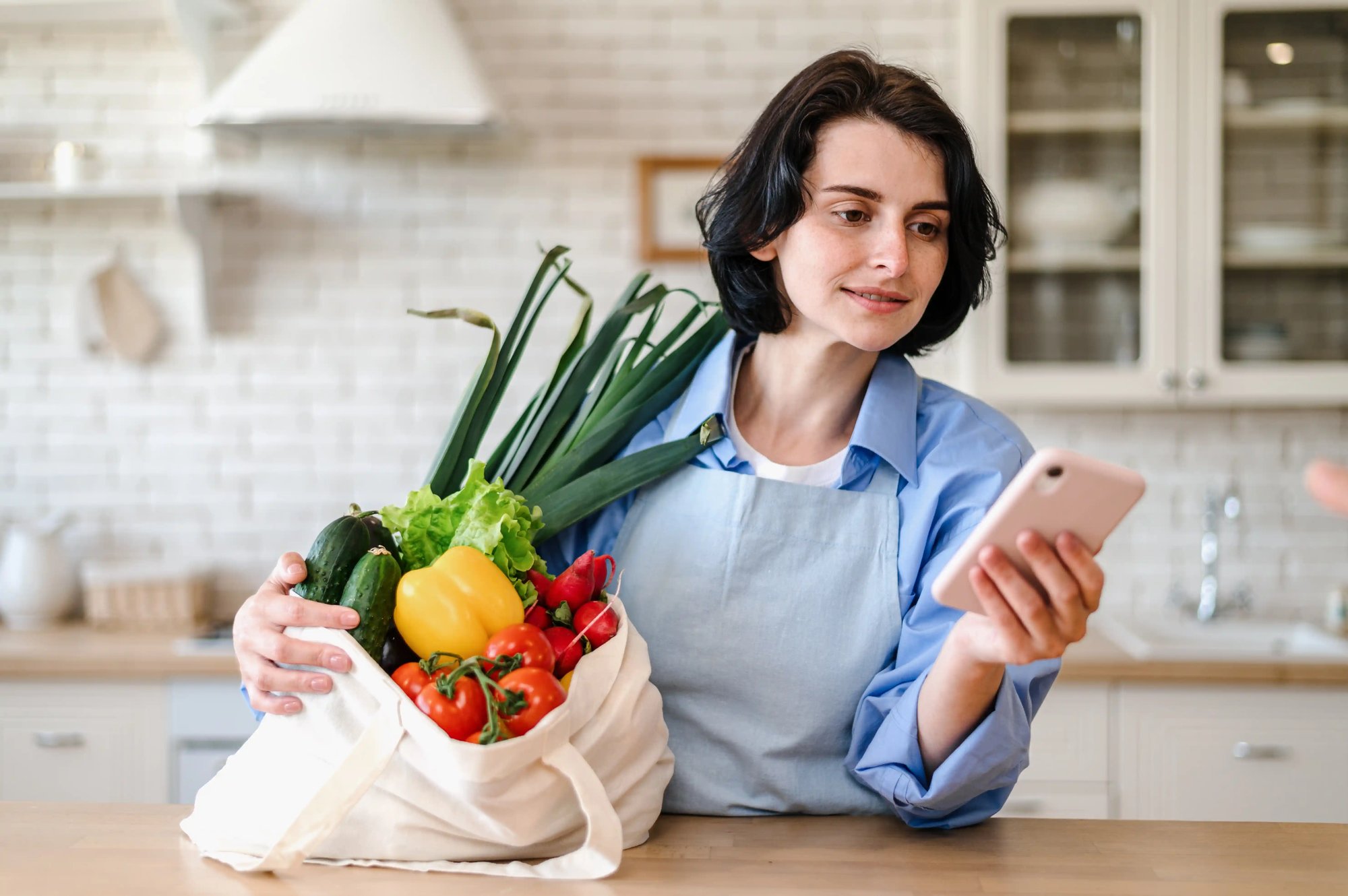 Mulher numa cozinha a segurar um saco reutilizável com legumes frescos enquanto consulta o telemóvel.