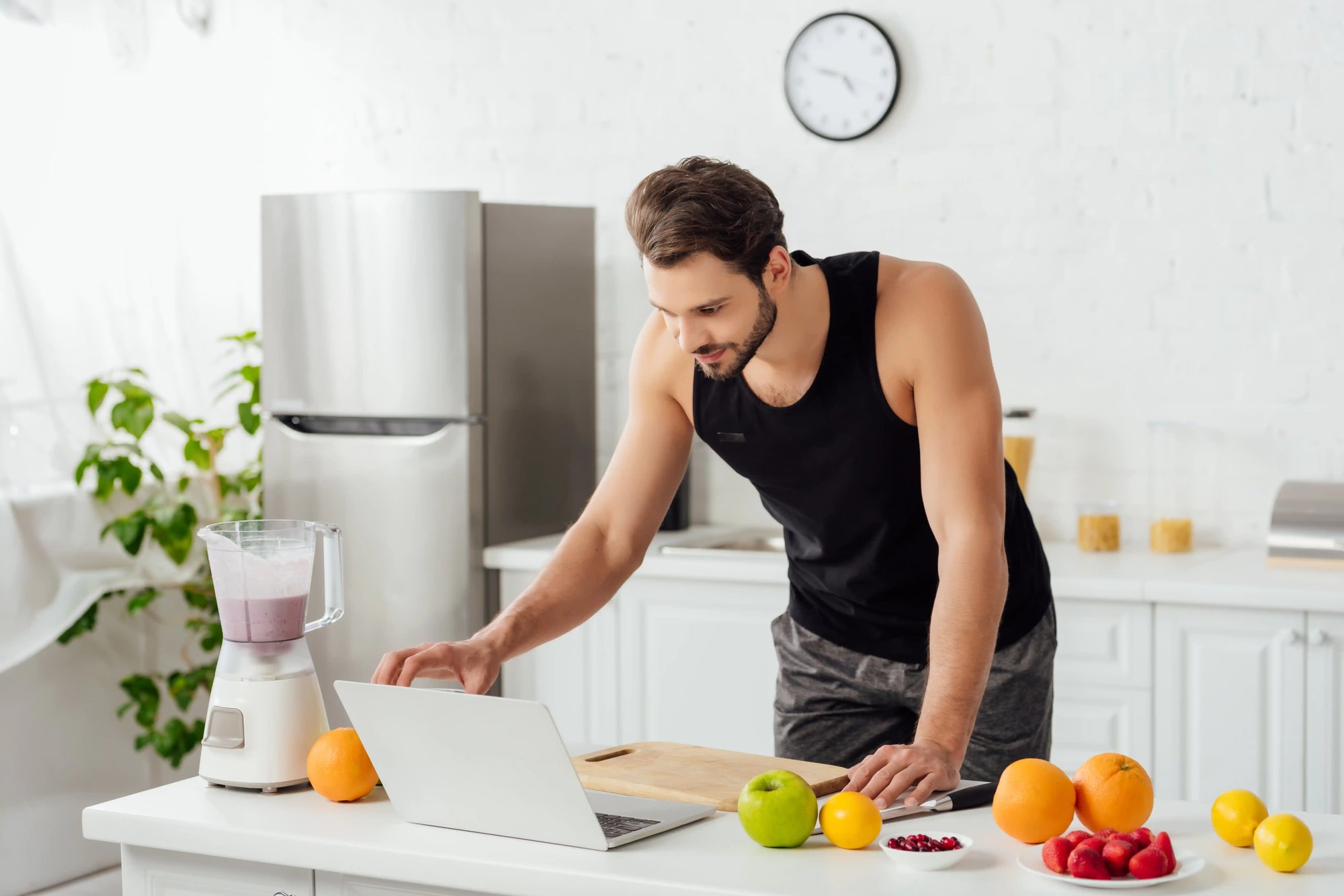 Homem numa cozinha a consultar um computador portátil junto a fruta fresca e um liquidificador com batido.