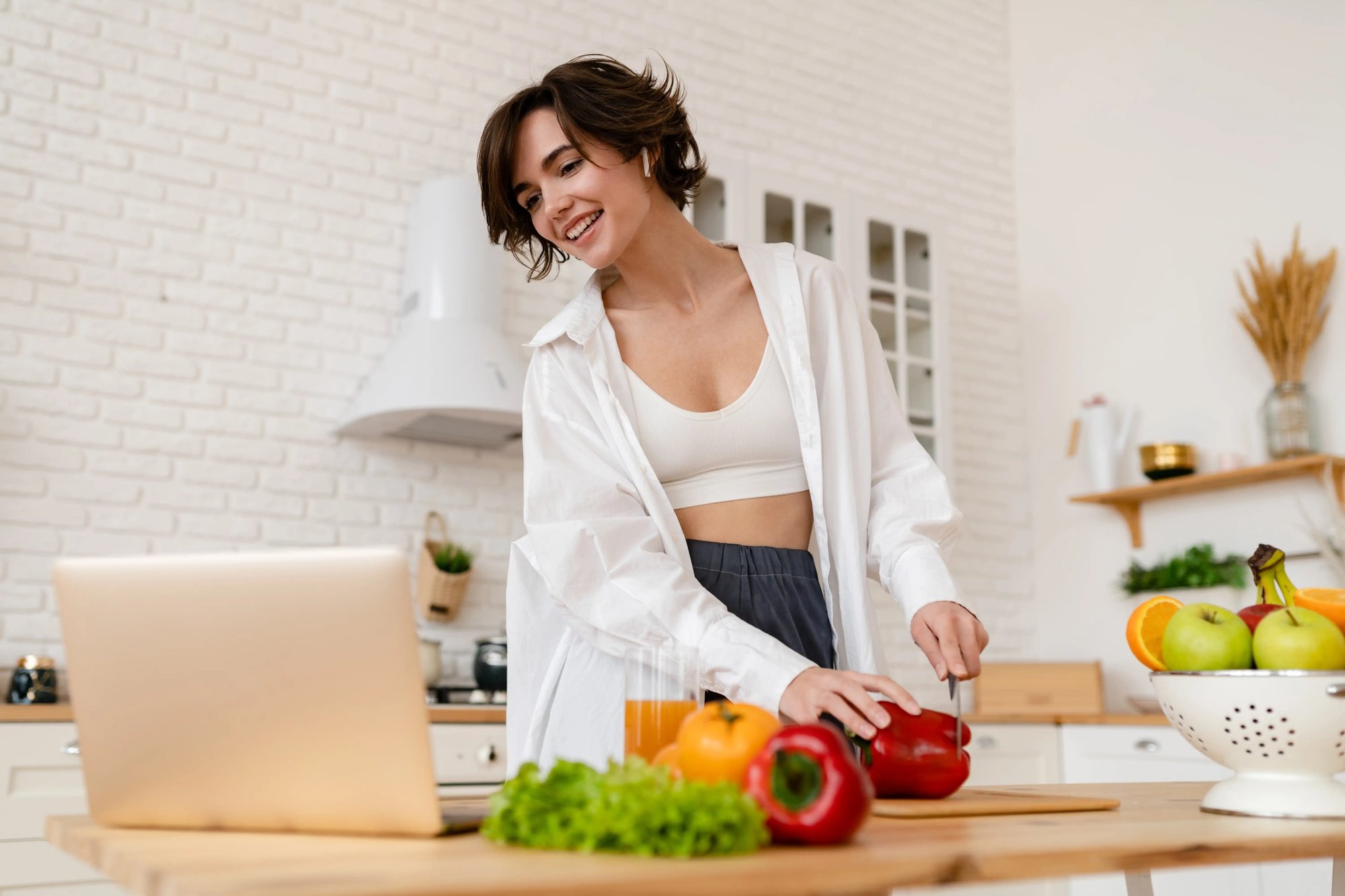 Mulher numa cozinha a preparar legumes frescos enquanto acompanha conteúdos num computador portátil.Mulher numa cozinha a preparar legumes frescos enquanto acompanha conteúdos num computador portátil.